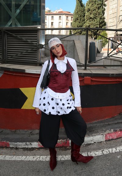 Top de capas de lunares rojos y blancos con un cuerpo ajustado burdeos, pantalones de pierna ancha negros y botas burdeos hasta la rodilla. Pañuelo de cabeza gris con patrón.