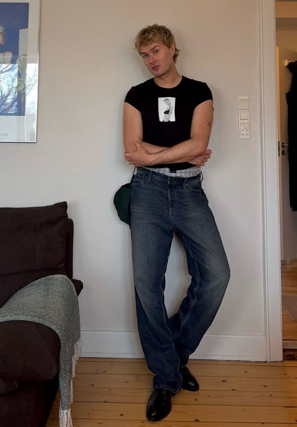 Person steht drinnen vor einer weißen Wand mit verschränkten Armen, trägt ein schwarzes T-Shirt mit einem monochromen Foto und lockeren blauen Jeans.