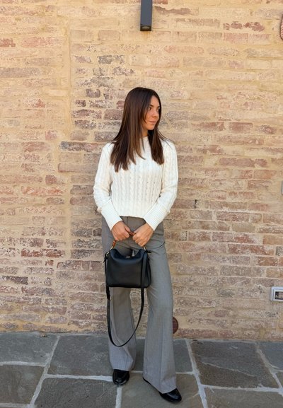 Suéter de punto con trenzas en color crema claro, pantalones acampanados grises y un pequeño bolso negro con acentos dorados. Fondo de pared de piedra.