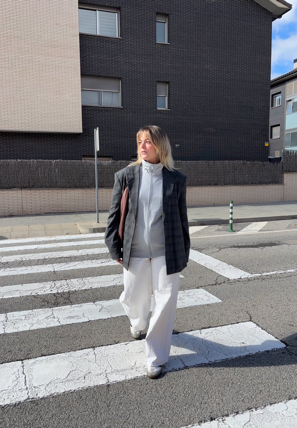 Persona con un ampio blazer a quadri grigio, una giacca bianca con zip e pantaloni bianchi a gamba larga, che cammina su un attraversamento pedonale con una pavimentazione testurizzata.
