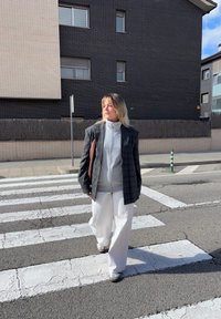 Persona con un ampio blazer a quadri grigio, una giacca bianca con zip e pantaloni bianchi a gamba larga, che cammina su un attraversamento pedonale con una pavimentazione testurizzata.