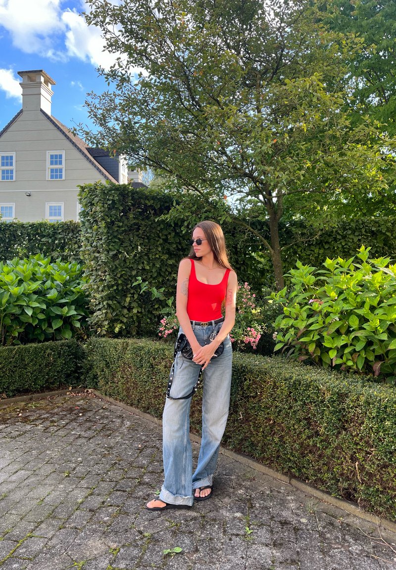 Κόκκινο αμάνικο τοπ, ανοιχτό μπλε φαρδιές παντελόνες με ραμμένες άκρες, μαύρη τσάντα μέσης με αλυσίδα και μαύρες σαγιονάρες σε εξωτερικό περιβάλλον.