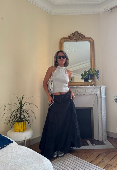 Top halter plateado, falda maxi negra con volantes y zapatillas plateadas. Los accesorios incluyen un bolso metálico. Entorno interior con plantas y un espejo.