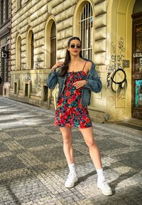 Λουλουδένιο φόρεμα με κόκκινες, ροζ και πράσινες σχέσεις; συνδυασμένο με τζιν μπουφάν και λευκά αθλητικά παπούτσια. Ο περιβάλλων χώρος διαθέτει υφή από ψηφίδα.