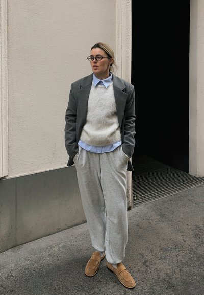 Blazer gris oversize sobre un suéter claro, con una camisa azul con cuello y pantalones deportivos grises. Zapatos deslizados color camel. Texturas suaves, ajuste relajado.