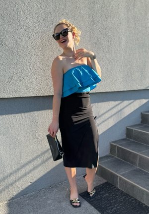 Falda negra ajustada con un top bandeau azul con volantes. Los accesorios incluyen gafas de sol negras y un clutch negro. Lleva sandalias negras sin talón con acentos.