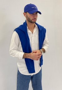 Modrá čiapka s logom, biela košeľa na gombíky, modrý sveter prehodený cez ramená a svetlo modré džínsy. Hladké textúry a voľný strih.