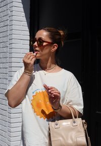 Ανοιχτόχρωμο oversized t-shirt με γραφικό ήλιου. Το άτομο κρατάει ένα ροζ προϊόν χειλιών και μια μπεζ τσάντα με μικρές μεταλλικές λεπτομέρειες. Φοράει γυαλιά ηλίου.