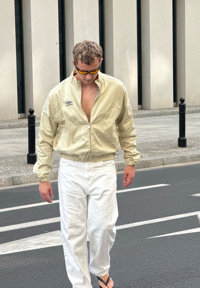 Homme portant une veste beige, un pantalon blanc, des tongs et des lunettes de soleil orange marchant dans une rue urbaine avec un trottoir en béton et des bornes noires.