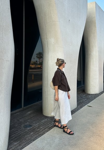 Camisa marrón de manga corta, falda blanca de pierna ancha, sandalias negras de tiras. Pared de hormigón lisa y curvada al fondo, suelo de madera.