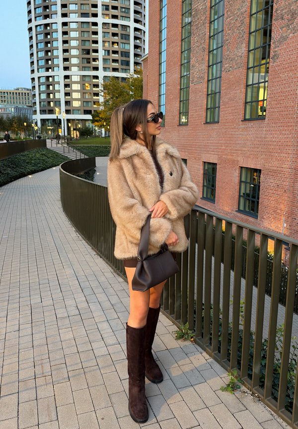 Una mujer con gafas de sol, un abrigo de piel beige y botas altas marrones está de pie junto a una barandilla entre edificios urbanos modernos.