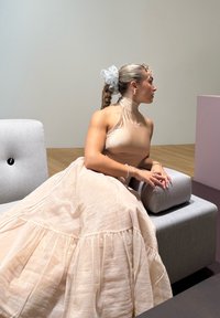 Vestido comprido até ao tornozelo, fluido e cor rosa claro, com uma barra franzida e um topo halter justo e brilhante. Assentos estofados em cinza suave ao fundo.