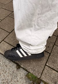 Zapatilla negra con rayas blancas usada con pantalones blancos, de pie sobre una acera pavimentada con musgo en las grietas.