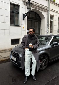 Sivi plašč, črn pulover, svetle hlače in črni škornji. V roki drži skodelico kave. Parkiran siv avto na mokri cesti, v ozadju stavba.