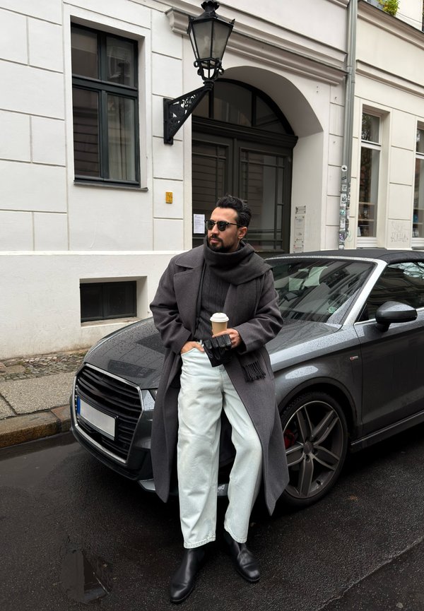 Grijze overjas, zwarte trui, lichtgekleurde broek en zwarte laarzen. Houdt een koffiekop vast. Grijze auto geparkeerd op natte straat, met gebouw op de achtergrond.