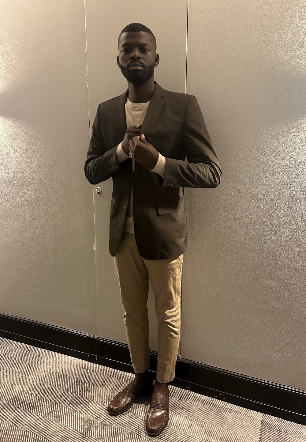 Homme ajustant un blazer marron foncé, portant un pantalon beige et des mocassins marron, debout devant un mur neutre sur un tapis à motifs.