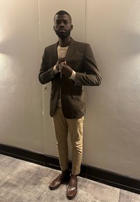 Homme ajustant un blazer marron foncé, portant un pantalon beige et des mocassins marron, debout devant un mur neutre sur un tapis à motifs.