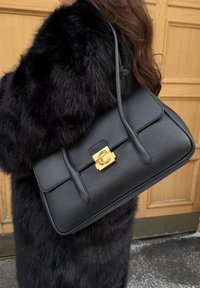 Femme portant un manteau en fausse fourrure noire, tenant un sac à main en cuir noir avec une fermeture dorée et deux bretelles épaules.