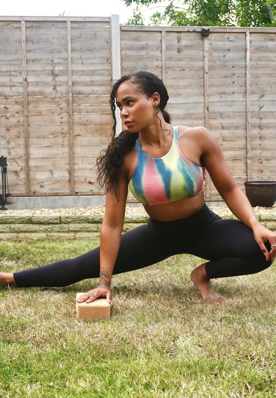 Sujetador deportivo con patrones de tie-dye en rosa, verde y azul; leggings negros de cintura alta; practicando yoga con un ladrillo sobre la hierba.