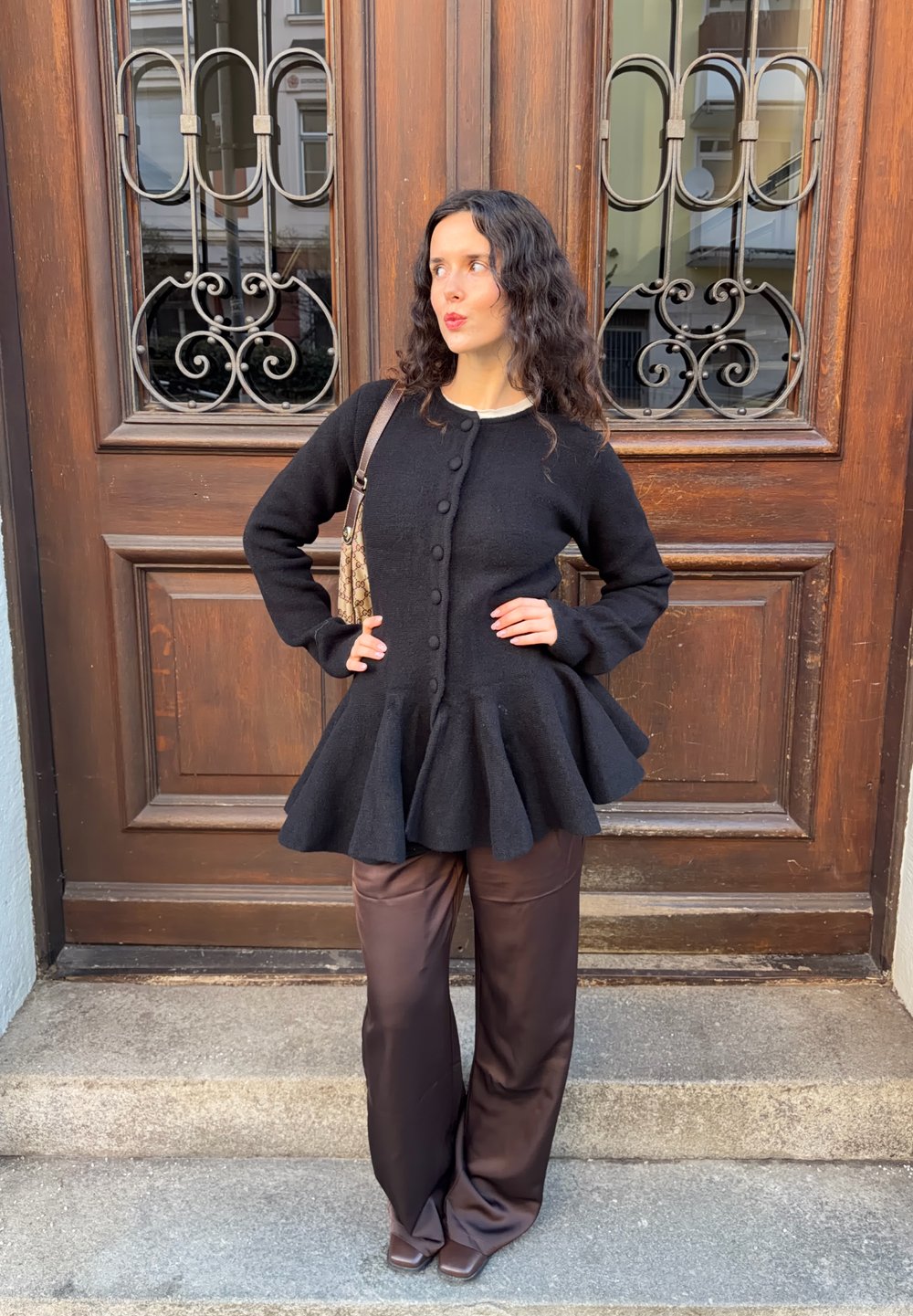 Pull noir à basque avec boutons, associé à un pantalon en satin marron. En arrière-plan, une porte en bois ornée de ferronnerie décorative.