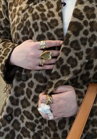 Καπαρντίνα με λεοπάρ σχεδίαση σε καφέ και μπεζ αποχρώσεις. Πολλοί χρυσοί δακτύλιοι με διάφορα σχέδια, συμπεριλαμβανομένων καρδιάς και ανθοσχημάτων, φορεμένοι στα δάχτυλα.