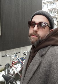 Homme barbu portant des lunettes de soleil, un bonnet gris, un sweat à capuche marron et un manteau gris debout à l'extérieur près de vélos recouverts de neige.