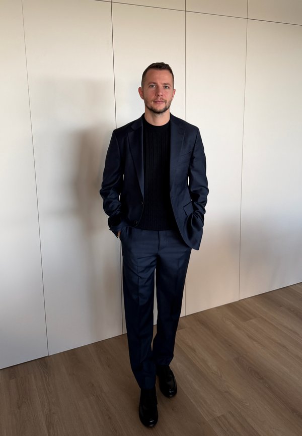 Hombre con traje azul oscuro y suéter negro, de pie con las manos en los bolsillos frente a una pared blanca con paneles sobre un suelo de madera clara.