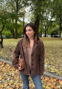 Brązowa zamszowa marynarka z sześcioma guzikami, nałożona na beżową bluzkę. Trzyma brązową plecioną torebkę. Otoczona jesiennymi liśćmi.