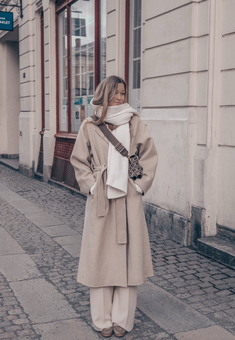 Femme portant un long manteau beige, une écharpe blanche et un sac bandoulière, debout sur une rue pavée près d'un bâtiment avec de grandes fenêtres.