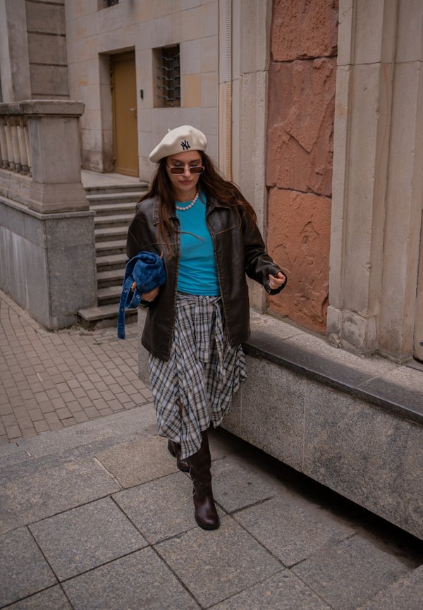 Mujer con boina beige, gafas de sol, top azul, falda de cuadros, chaqueta de cuero negra y botas marrones caminando por una acera urbana de piedra.