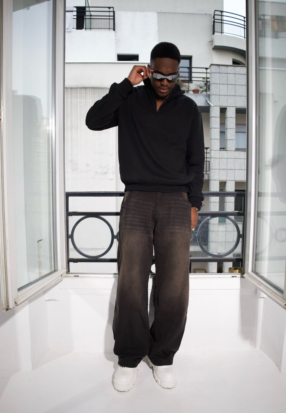 Pull noir avec un col montant, associé à un pantalon gris foncé ample et des baskets blanches au look épais. Les accessoires incluent des lunettes de soleil.
