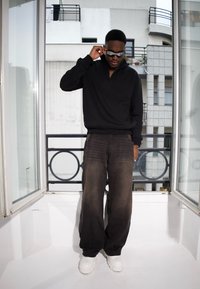 Pull noir avec un col montant, associé à un pantalon gris foncé ample et des baskets blanches au look épais. Les accessoires incluent des lunettes de soleil.