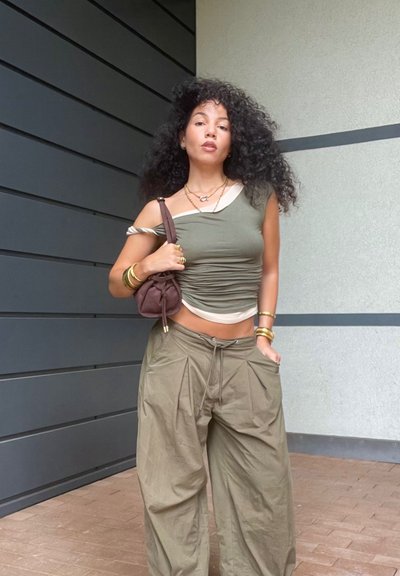 Mujer con cabello rizado, vistiendo un top corto arrugado color oliva y pantalones de pierna ancha, sosteniendo un bolso marrón cruzado, de pie contra las paredes de un edificio moderno.