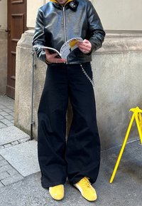 Μαύρο δερμάτινο μπουφάν, κίτρινη φούτερ από κάτω, φαρδιά μαύρα παντελόνια, κίτρινες αθλητικές μπότες. Κρατάει ένα περιοδικό, ασημί λεπτομέρεια στην παντελόνι.