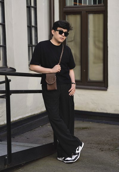 Joven con camisa negra y pantalones a rayas se apoya en la barandilla de un edificio, lleva gafas de sol, zapatillas deportivas y un bolso bandolera marrón.