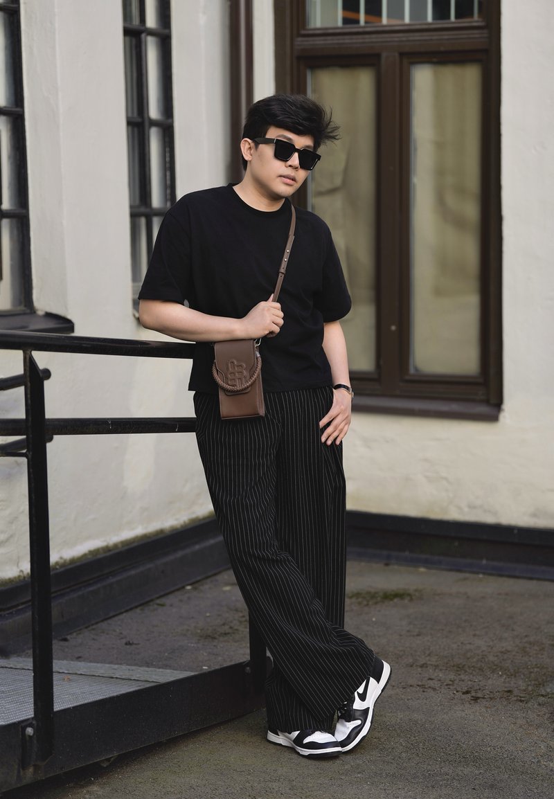 Jeune homme en chemise noire et pantalon rayé appuyé sur une rambarde devant un bâtiment, portant des lunettes de soleil, des baskets et un sac bandoulière marron.