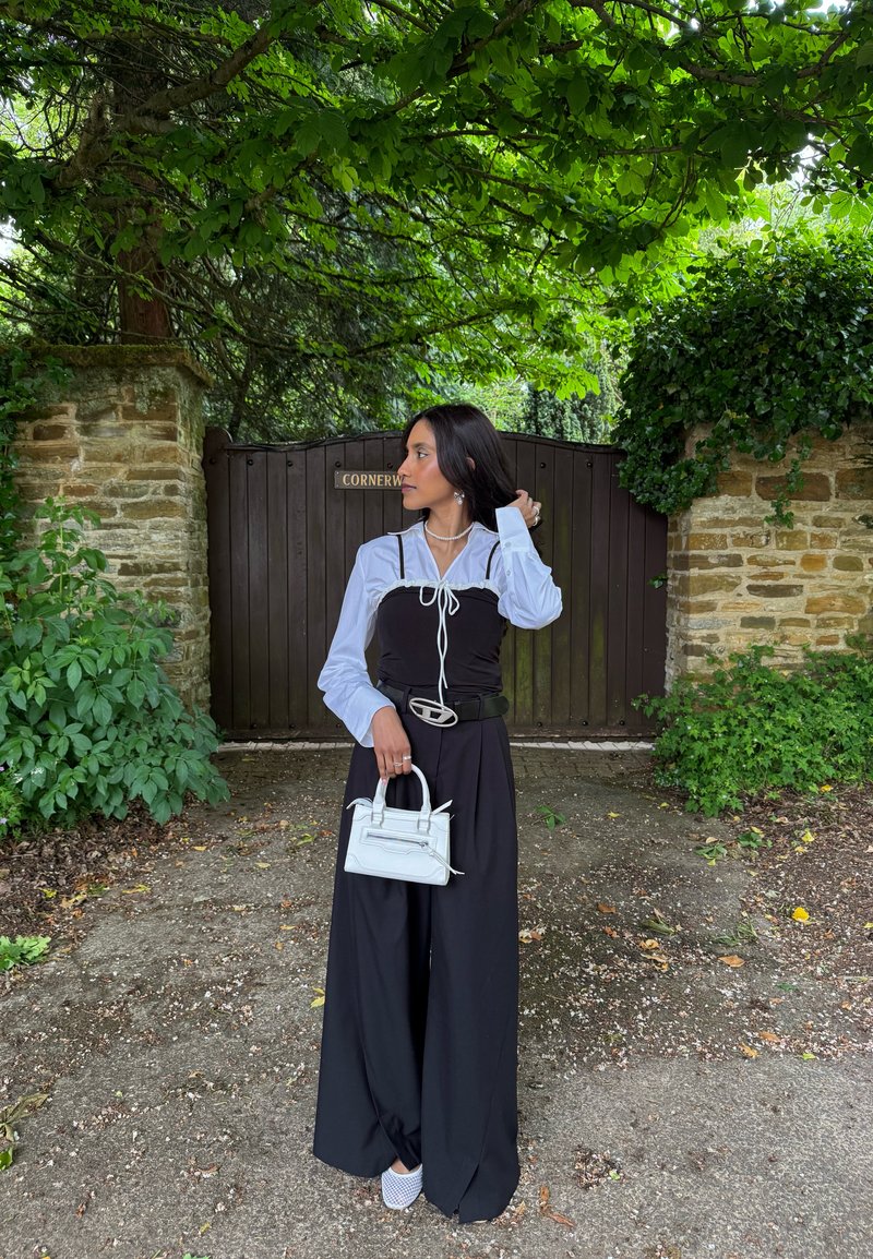 Femme en pantalon noir large, chemise bleu clair et sac blanc, debout sur un sentier devant une porte en bois entourée de verdure.