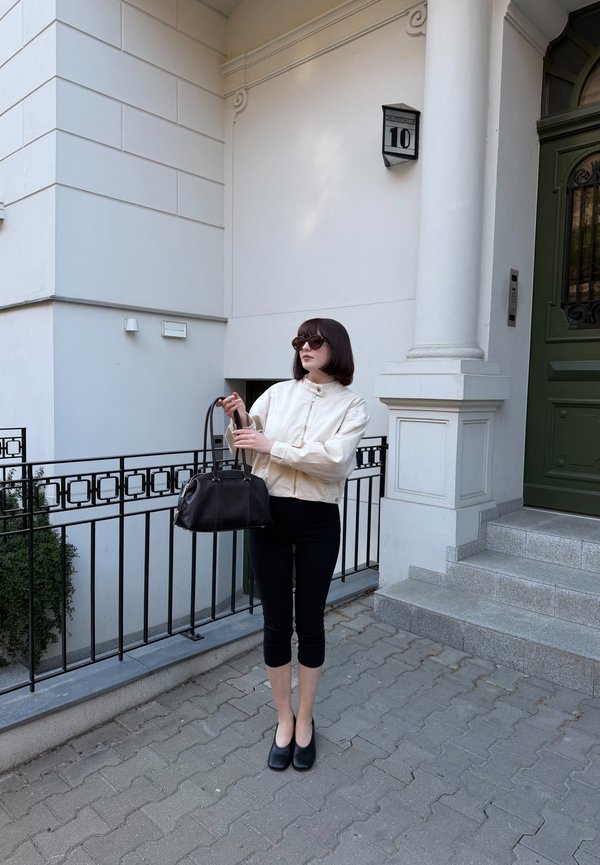 Kvinde iført solbriller, beige jakke og sorte korte bukser holder en sort håndtaske og står på en belagt fortov ved hvide bygnings-trin.