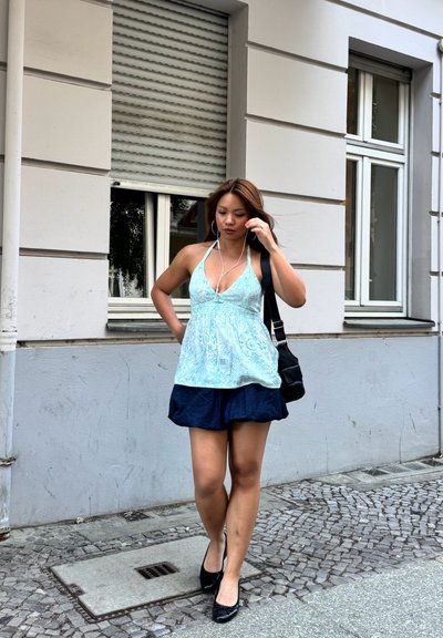 Mujer caminando por una acera de adoquines con top azul sin mangas, falda azul oscuro, zapatos negros, llevando un bolso negro, con auriculares en los oídos.