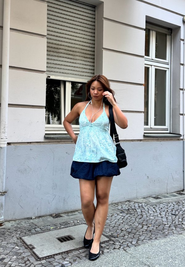 Žena kráča po dláždenom chodníku, oblečená v modrý tielkový top, tmavomodrú sukňu, čierne topánky, nesie čiernu tašku, s slúchadlami v ušiach.