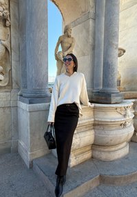 Femme portant des lunettes de soleil, un pull blanc et une jupe noire, appuyée contre une structure en marbre ornée avec une statue classique en plein air.