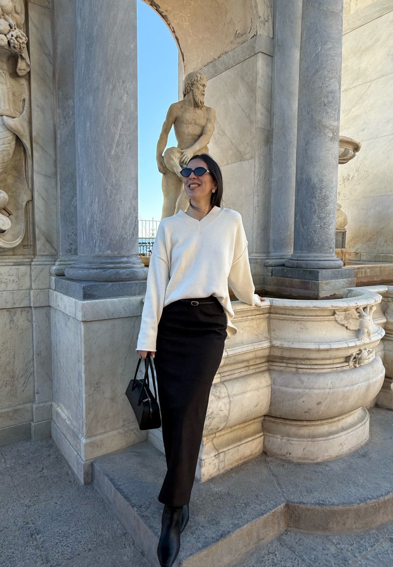 Femme portant des lunettes de soleil, un pull blanc et une jupe noire, appuyée contre une structure en marbre ornée avec une statue classique en plein air.