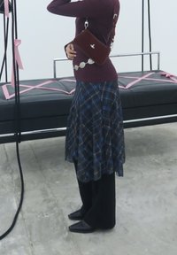A person in a maroon top, wearing a plaid skirt over black trousers and black shoes, stands next to a black padded swing seat with pink straps indoors.