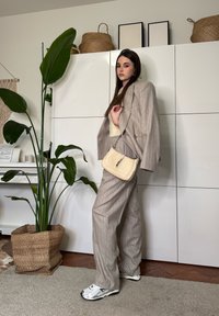 Femme en tailleur beige à fines rayures et baskets tenant un sac à main crème, debout à l'intérieur près d'une grande plante verte et d'une armoire blanche avec des paniers tissés.