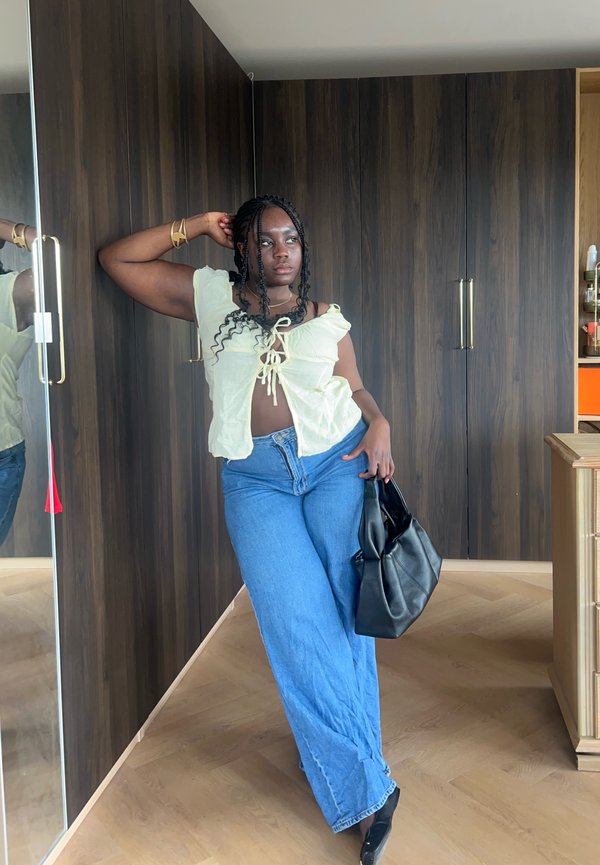 Femme aux cheveux tressés portant un haut jaune pâle et un jean bleu, tenant un sac à main noir, appuyée contre une armoire en bois foncé.