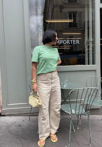 Grün-weiß gestreiftes, cropped T-Shirt, beiges, weites Hosen, gelbe Flip-Flops und eine kleine hellgelbe Handtasche. Außencafé-Setting.
