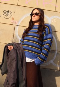 Pull en maille rayé bleu et noir avec des accents jaunes ; le modèle tient une veste sombre, debout devant un mur couvert de graffitis.