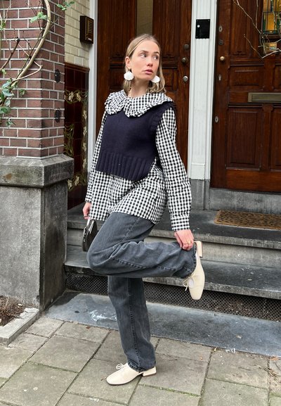 Chaleco de punto azul marino sobre una camisa de cuadros en blanco y negro, combinado con jeans grises y zapatos planos crema. Los accesorios incluyen un pequeño bolso negro.