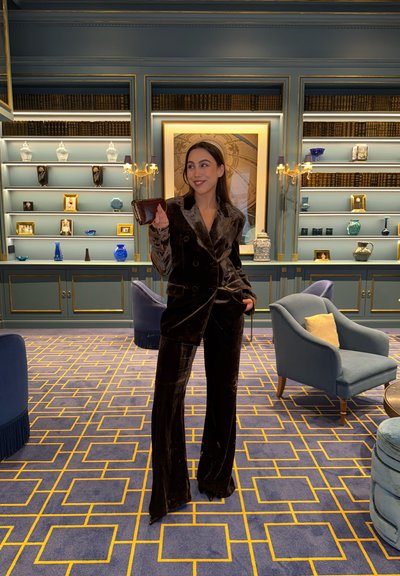 Mujer con traje de terciopelo oscuro sosteniendo un bolso pequeño, de pie en una habitación alfombrada con un patrón azul y dorado, con estanterías y sillones.