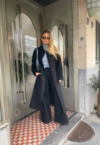 Casaco preto longo sobre um suéter de gola alta azul claro, emparelhado com calças pretas de perna larga. Óculos de sol elegantes e uma bolsa cinza completam o visual.